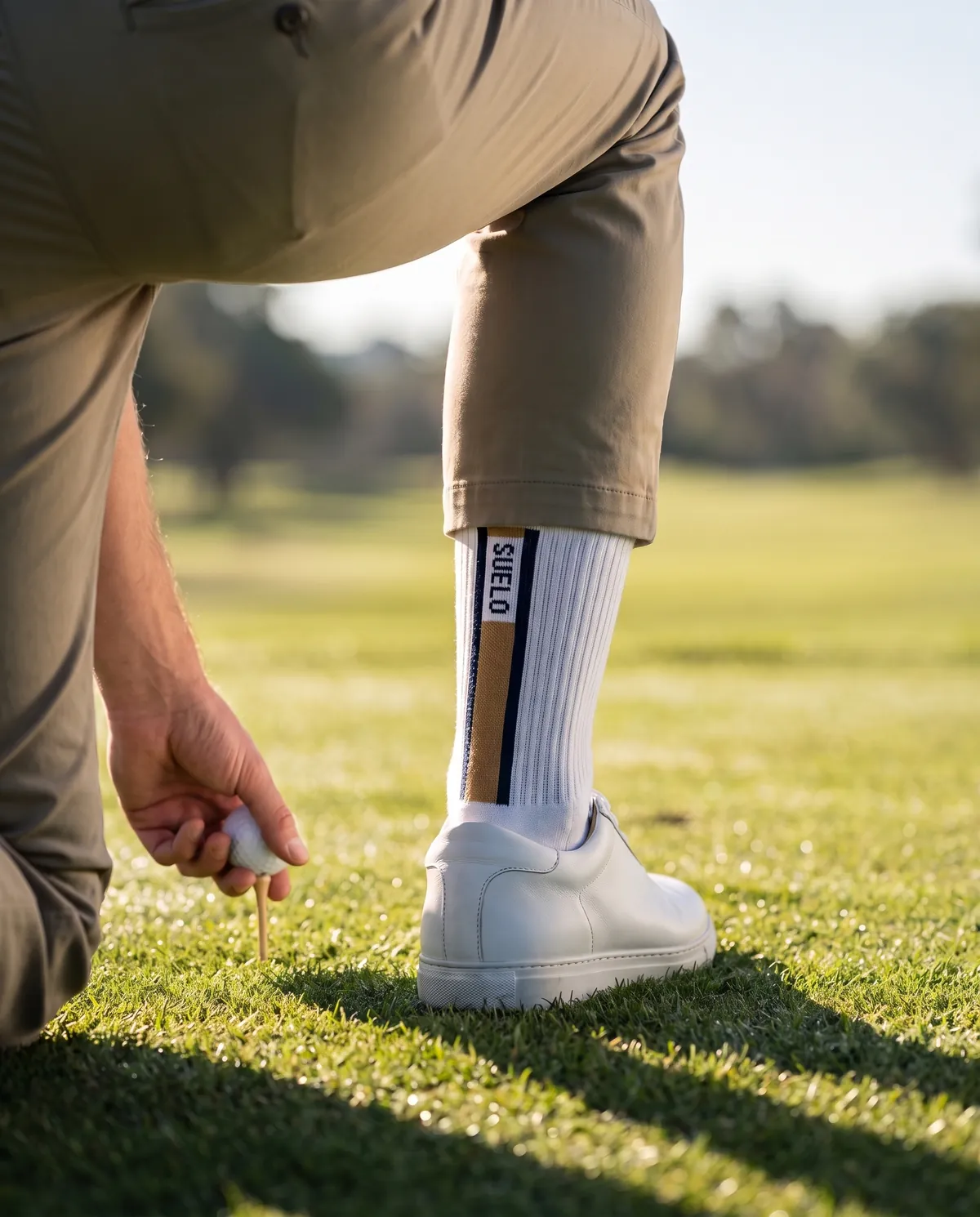 A golfer tees up a ball, the Suelo stripe visible on the back of the sock above the shoe.